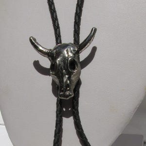 May include: A bolo tie featuring a silver-toned metal steer skull pendant centred on a black braided leather cord. The cord ends are finished with matching silver-toned metal tips that have a ribbed, cylindrical design for a secure hold.