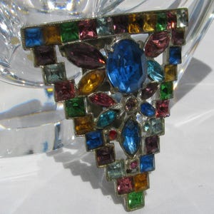 May include: A triangular brooch featuring a silver-tone metal frame encrusted with multi-coloured faceted rhinestones. The design includes a large central oval blue stone surrounded by smaller square, marquise, and round stones in shades of blue, green, purple, pink, and amber.