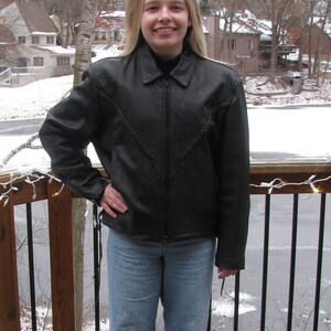 May include: A woman wearing a black leather jacket with a braided design, blue jeans, and black shoes. She is standing on a wooden deck with a railing.