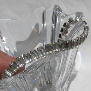May include: A silver-toned bracelet with rectangular, clear crystals. The bracelet is resting on the edge of a clear, faceted glass bowl. The crystals are set closely together, creating a continuous line of sparkle. The bowl has a starburst design.