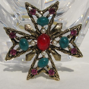 Puede incluir: Broche de cruz dorada vintage con una piedra ovalada roja central, rodeada de piedras turquesas y magenta, y peque&ntilde;as perlas de imitaci&oacute;n. El broche tiene un dise&ntilde;o detallado y texturizado.