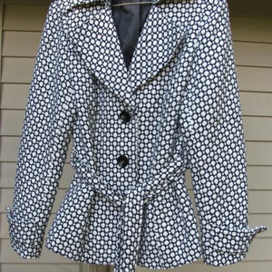 May include: A black and white patterned jacket with a tie belt. The jacket has a notched collar, long sleeves, and three black buttons down the front. The pattern is a repeating design of small white circles on a black background.