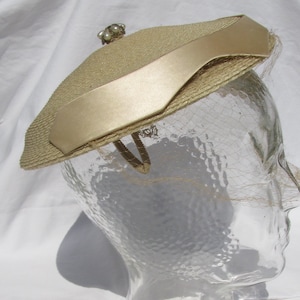 May include: A beige straw hat with a smooth satin ribbon band wrapped around the crown. The hat features a small cluster of white faux pearls at the top center and a fine, delicate netting veil that extends downward from the brim edge.