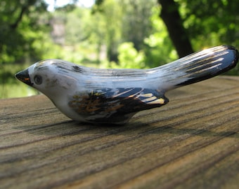 以下が含まれることがあります： 黒と金のアクセントが付いた小さな白い磁器製の鳥の置物。鳥は木製の表面に置かれています。