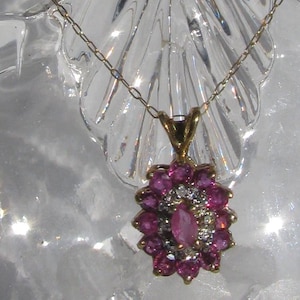 May include: A gold pendant necklace featuring a cluster of pink gemstones and small diamonds. The pendant is flower-shaped, with a central oval pink stone. The necklace is displayed on a clear, decorative glass object.