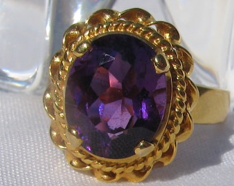 Puede incluir: Un anillo de oro con una gran amatista ovalada. El anillo tiene un diseño detallado, como de cuerda, alrededor de la piedra.