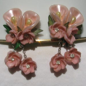 May include: A pair of clip-on earrings featuring two pale pink calla lily shapes with cream-colored centers, accented by small pink flowers with green leaves. Each earring has two additional pink flower charms dangling from silver-toned metal chains.