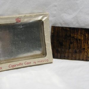 May include: A rectangular cigarette case by Mavco with a dark brown and black tortoiseshell pattern, featuring a ribbed texture. It is presented in its original off-white cardboard box with thin horizontal stripes and a clear plastic window.