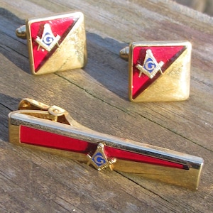 May include: A set of gold-tone Masonic cufflinks and tie bar. The cufflinks and tie bar feature a red and gold enamel design with a compass and square.