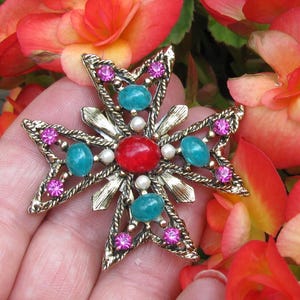 Puede incluir: Broche dorada en forma de cruz con una piedra central roja, detalles turquesa y rosa, y perlas de imitaci&oacute;n. La broche se encuentra sobre un fondo de flores rojas y naranjas vibrantes.
