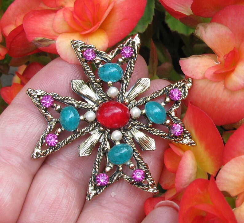 Puede incluir: Broche dorada en forma de cruz con una piedra central roja, detalles turquesa y rosa, y perlas de imitaci&oacute;n. La broche se encuentra sobre un fondo de flores rojas y naranjas vibrantes.