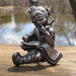 May include: A silver-toned figurine of a seated, bespectacled figure reading a book. The figure is wearing a robe and has a thoughtful expression, with a finger touching their lips. The figurine is on a wooden surface.