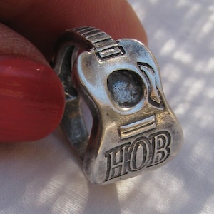 May include: Silver guitar ring with the word "HOB" engraved on the body. The ring is designed to look like a real guitar.