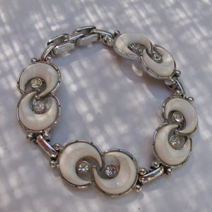 May include: A silver-tone link bracelet featuring repeating circular motifs. Each motif contains two white crescent-shaped cabochons and a central clear round rhinestone. The bracelet is constructed with polished metal links connecting the decorative segments.