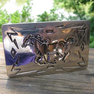 May include: A rectangular silver-colored belt buckle with a detailed relief of a running horse. The buckle has decorative geometric patterns and lightning bolt designs around the edges. The horse is the central focus.