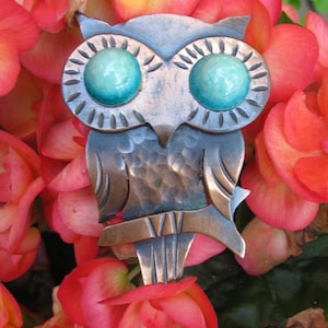 May include: A metal owl brooch with turquoise-colored eyes and a textured, bronze-colored body. The owl is set against a backdrop of vibrant red and orange flowers, creating a striking contrast. The brooch is a decorative accessory.