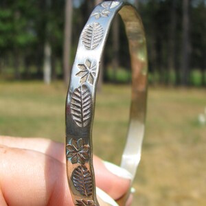 May include: A silver-toned metal bangle bracelet featuring a repeating embossed pattern of small leaves and star-shaped flowers along its outer surface. The bracelet has a smooth, polished finish and a slightly irregular, organic shape.