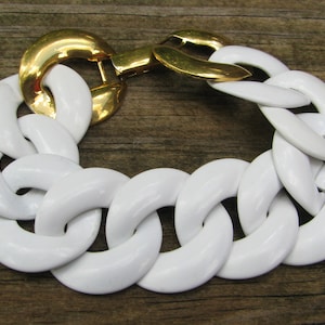 Pode incluir: Uma pulseira de elos brancos grossos com fecho dourado. A pulseira apresenta elos brancos grandes e brilhantes conectados por um fecho dourado. A joia está sobre um fundo de madeira.