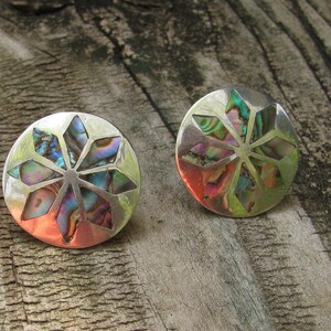 Puede incluir: Un par de pendientes de plata con un dise&ntilde;o circular que presenta una incrustaci&oacute;n de n&aacute;car de abul&oacute;n multicolor. La incrustaci&oacute;n es un patr&oacute;n de estrella con un punto central.