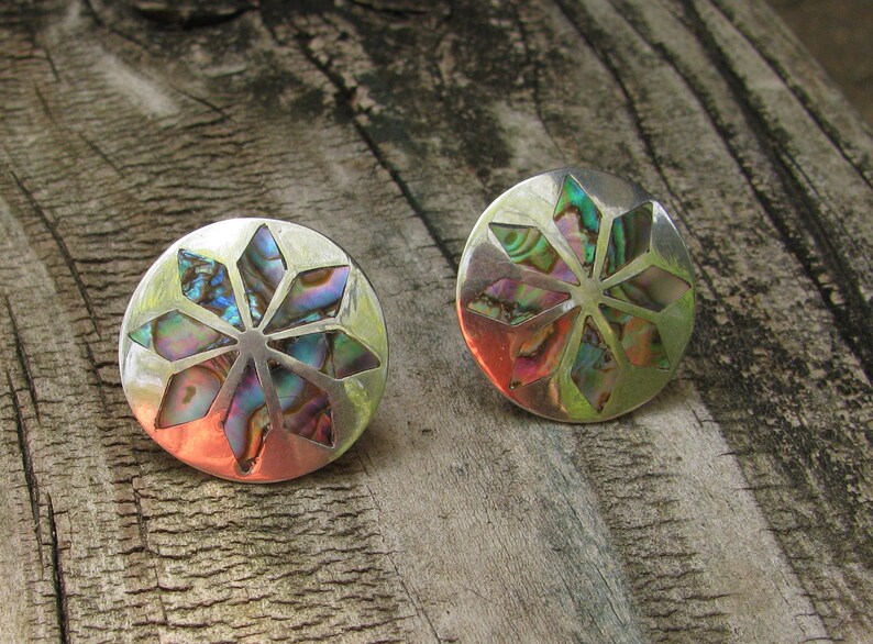 Puede incluir: Un par de pendientes de plata con un dise&ntilde;o circular que presenta una incrustaci&oacute;n de n&aacute;car de abul&oacute;n multicolor. La incrustaci&oacute;n es un patr&oacute;n de estrella con un punto central.