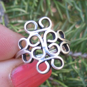 May include: A silver-toned ring with an openwork design featuring a series of interconnected, swirling shapes. The ring's design is centered around a cluster of loops and curves, creating a decorative and intricate pattern. The ring is shown against a blurred green background.
