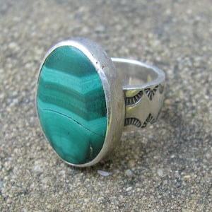 May include: A silver ring featuring a large, oval-shaped green malachite gemstone with distinct banded patterns. The metal band is wide and decorated with stamped, fan-like geometric engravings along the sides of the setting.