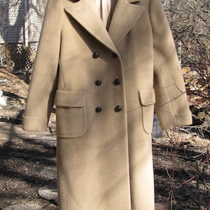 May include: A tan double-breasted coat with black buttons and two front pockets. The coat is hanging on a wooden hanger outdoors. The coat has a classic, tailored design.