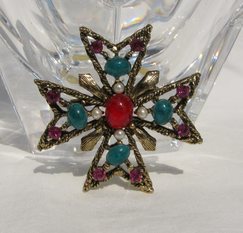 Puede incluir: Broche de cruz dorada vintage con una piedra ovalada roja central, rodeada de piedras turquesas y magenta, y peque&ntilde;as perlas de imitaci&oacute;n. El broche tiene un dise&ntilde;o detallado y texturizado.