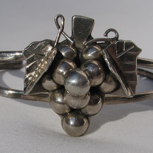 May include: A silver bracelet featuring a grape cluster design. The grapes are made of round beads and the leaves have a textured finish.