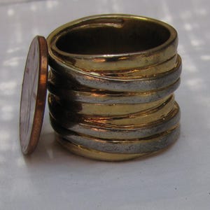 May include: A stack of gold and silver rings with a penny next to them. The rings have alternating bands of gold and silver metal. The penny is copper-coloured, providing a size comparison.
