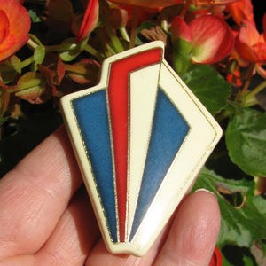 May include: A white ceramic brooch with a geometric design in red, blue, and gold. The brooch is shaped like a diamond and has a modern, abstract look.