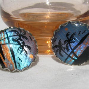 May include: A pair of round earrings with a silver-coloured scalloped edge. The earrings feature a design of palm trees and a sunset scene in shades of blue, orange, and black. The earrings are set against a soft white background.