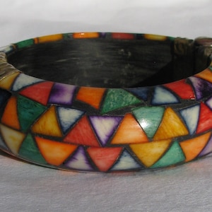 Può includere: Un braccialetto colorato con un design a mosaico di triangoli in tonalità di verde, blu, giallo, arancione, rosso e viola. Il braccialetto ha una fascia in metallo dorato intorno alla parte superiore.