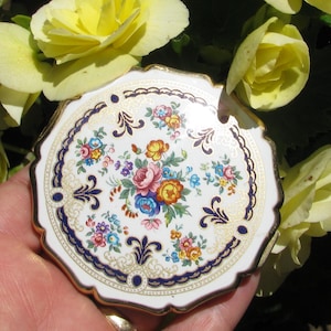 May include: A vintage, white, scalloped-edge compact with a floral design. The compact features a gold trim, a navy blue border, and a central bouquet of pink, yellow, blue, and orange flowers. The compact is held in a hand.