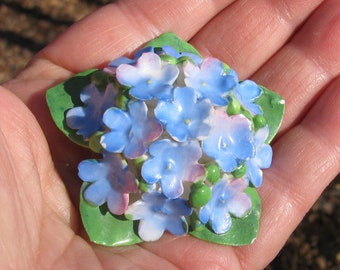 May include: A porcelain flower brooch with blue and white forget-me-not flowers and green leaves.