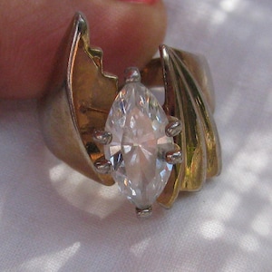 May include: A ring featuring a central marquise-cut clear gemstone held by four prongs. The band is constructed from two distinct metal sections, one with a smooth gold-tone finish and the other with a ribbed, layered gold-tone design on the side.