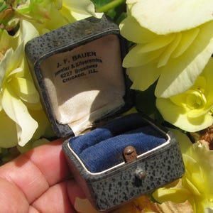 May include: A dark gray textured jewelry box with a hinged lid, featuring a blue velvet interior and a cream-colored satin lining printed with the text J. F. BAUER Jeweler & Silversmith 6227 Broadway CHICAGO, ILL. The box has a metal latch.