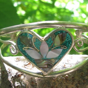 以下が含まれることがあります： ハート型のチャームが付いたシルバーブレスレット。チャームはターコイズとアワビの貝殻のインレイで飾られており、スタイリッシュな花のデザインになっています。