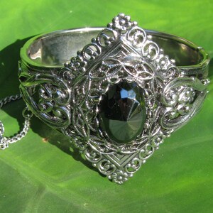 May include: A silver bracelet with an ornate design and a large black gemstone in the center.