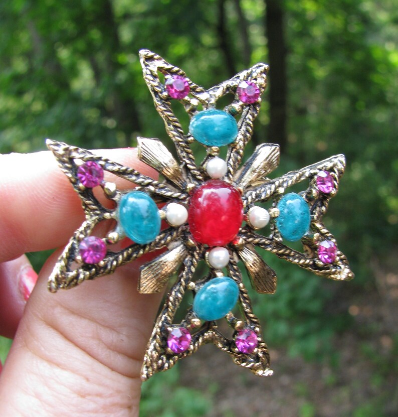 Puede incluir: Broche vintage en forma de cruz dorada. El broche presenta una piedra roja central, rodeada de piedras preciosas turquesas y rosas, y peque&ntilde;as perlas blancas. El dise&ntilde;o incluye intrincados trabajos en metal y un acabado texturizado, creando un accesorio ornamentado.