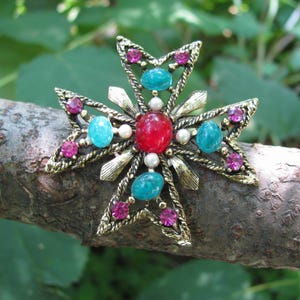 Puede incluir: Broche de estilo vintage en forma de cruz, con un marco de metal dorado. El broche est&aacute; adornado con piedras preciosas rojas, turquesas y rosas, junto con peque&ntilde;as perlas blancas. El fondo es natural, con una rama.