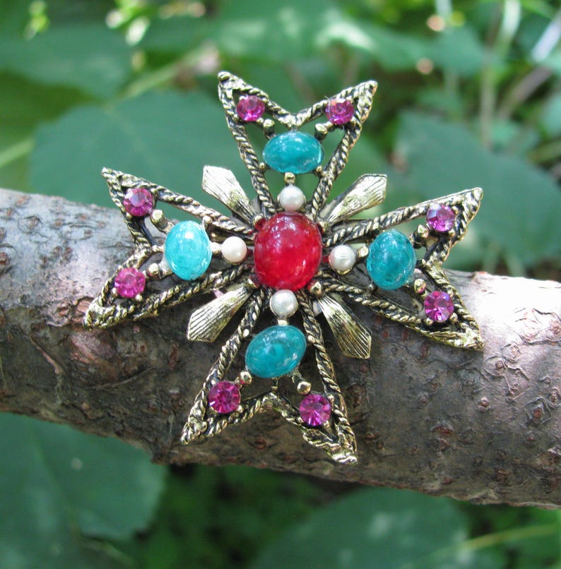 Puede incluir: Broche de estilo vintage en forma de cruz, con un marco de metal dorado. El broche est&aacute; adornado con piedras preciosas rojas, turquesas y rosas, junto con peque&ntilde;as perlas blancas. El fondo es natural, con una rama.