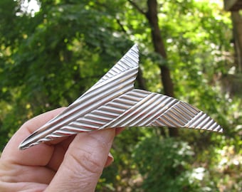 May include: A silver-toned, geometric brooch with a textured, striped design. The brooch is shaped like a sharp, pointed wing.