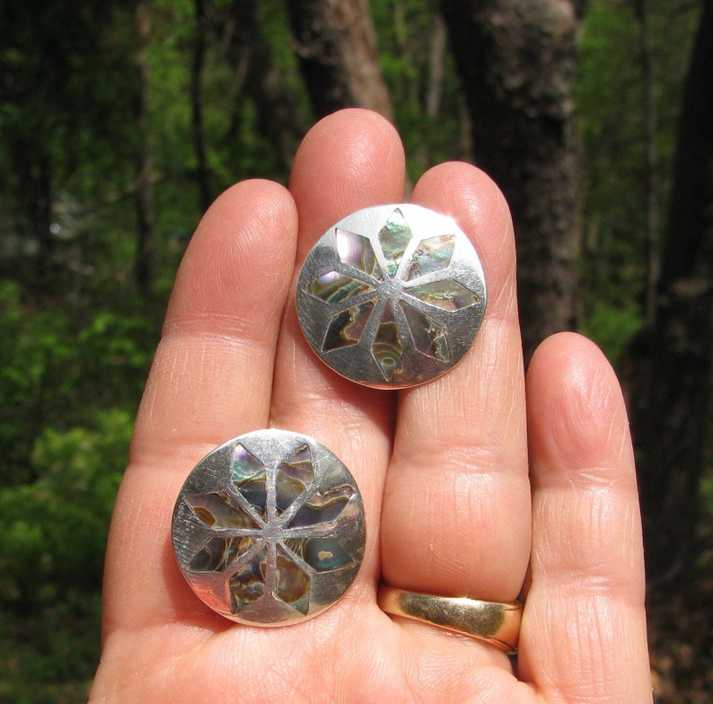 Puede incluir: Un par de pendientes de plata con un dise&ntilde;o circular de concha de abul&oacute;n. La concha tiene un patr&oacute;n geom&eacute;trico con una forma de estrella en el centro. Los pendientes son de color plata claro con un acabado brillante.
