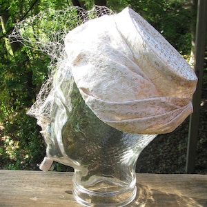May include: A white vintage hat with a lace design and a veil. The hat is on a clear glass display stand.