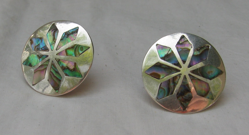 Puede incluir: Un par de pendientes de plata con un dise&ntilde;o circular que presenta un patr&oacute;n de estrella en forma de estrella hecho de concha de abul&oacute;n iridiscente.