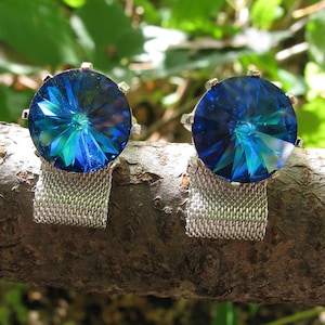 May include: Two cufflinks with large, round, faceted blue crystals. The crystals have a vibrant, multi-toned appearance and are set in silver-toned metal with a mesh backing. The cufflinks are displayed on a natural wooden branch.