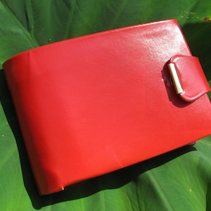 May include: A vibrant red wallet, rectangular in shape, with a clasp closure. The wallet is made of a smooth material and is set against a green leaf background.