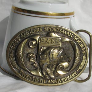 May include: A brass belt buckle with the words "Bass Anglers Sportsman Society 1967-1987 Twentieth Anniversary" and a bass fish design. The buckle is oval shaped and has a gold finish.