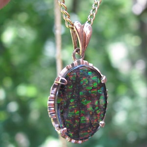 May include: A gold-toned necklace with an oval pendant. The pendant features a dark gemstone with a mosaic of green, red, and blue colors. The chain is a delicate link style, and the pendant has a decorative frame.